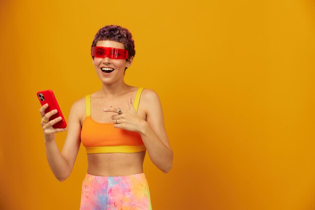 Mujer bloguera sorprendida con gafas milenarias inusuales tomando selfies en su teléfono luciendo ropa de colores brillantes contra un espacio libre de fondo de estudio naranja