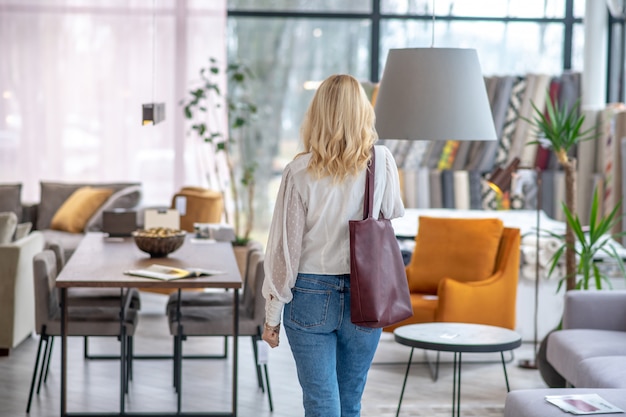 Mujer con una blusa blanca con una bolsa de color burdeos en el hombro, de pie con la espalda en el salón de muebles.