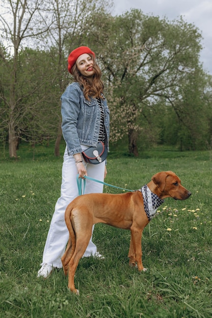 mujer en boina roja caminando con cachorro marrón de Rhodesian Ridgeback en campo verde en el parque