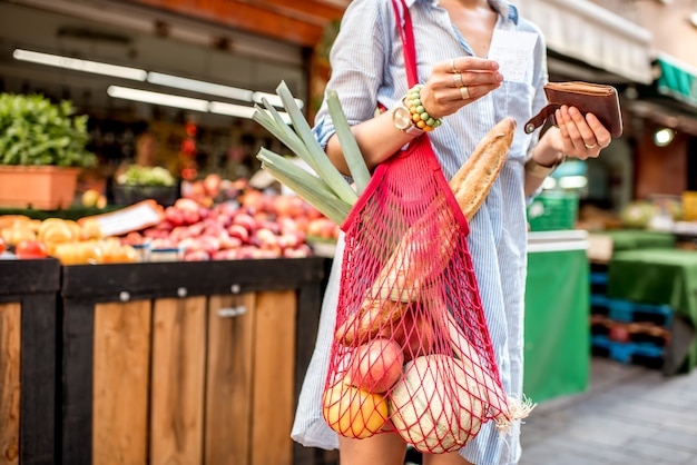 Mujer con bolsa de malla llena de verduras frescas mirando el cheque de compra en frente de la tienda de alimentos en Francia