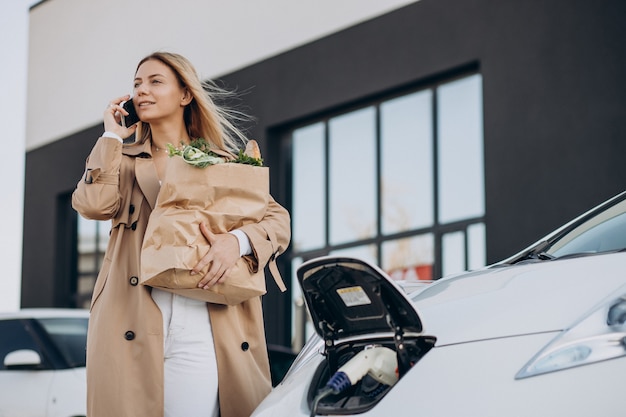 Mujer con bolsas de la compra de alimentos cargando coche eléctrico