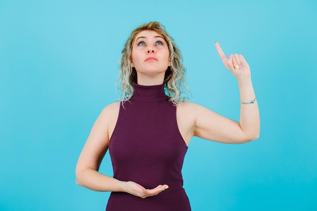 La mujer bonde está mirando y apuntando hacia arriba con el dedo índice sobre fondo azul.