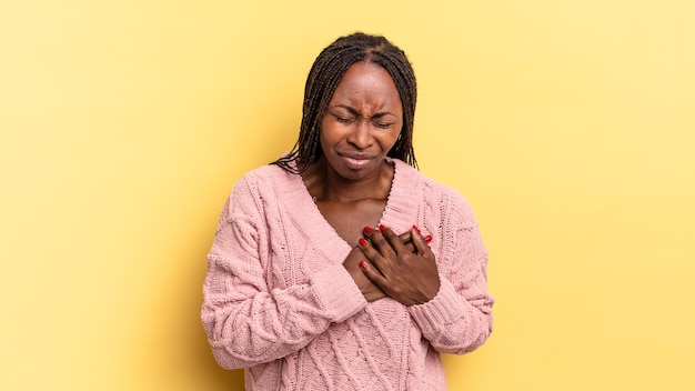 Mujer bonita afro negra que parece triste, herida y con el corazón roto, sosteniendo ambas manos cerca del corazón, llorando y sintiéndose deprimida