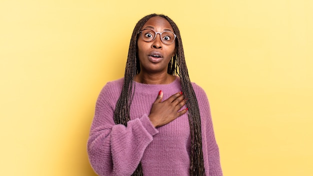 Mujer bonita afro negra que se siente conmocionada y sorprendida, sonriendo, tomando la mano al corazón, feliz de ser la indicada o mostrando gratitud