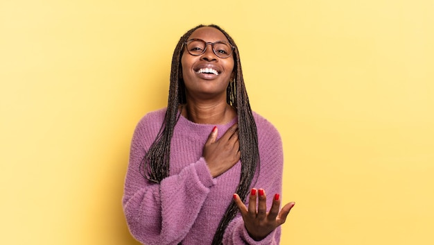 Mujer bonita afro negra que se siente feliz y enamorada, sonriendo con una mano al lado del corazón y la otra estirada al frente