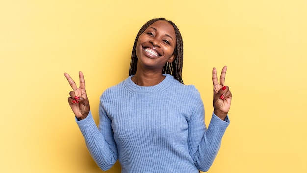 Mujer bonita afro negra sonriendo y luciendo feliz, amigable y satisfecha, gesticulando victoria o paz con ambas manos