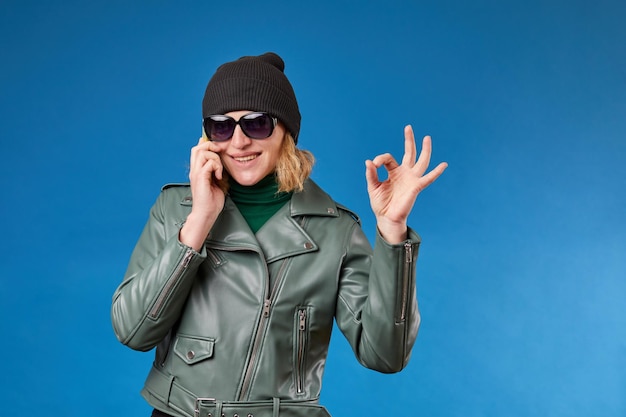 Una mujer bonita y alegre con una chaqueta verde brillante sostiene un teléfono inteligente en la mano y muestra el signo OK haciendo gestos con los dedos sonriendo lindamente mirando la cámara aislada sobre fondo azul con espacio publicitario para copiar