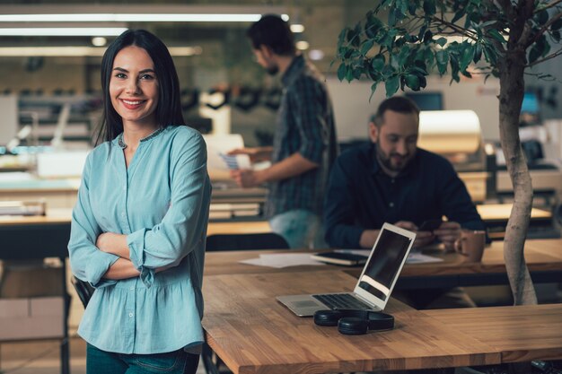 Mujer bonita alegre de pie junto a la mesa y sonriendo mientras sus colegas están en el fondo. Banner de plantilla