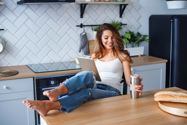 Mujer bonita alegre que usa la tableta y bebiendo el café en la cocina en casa.