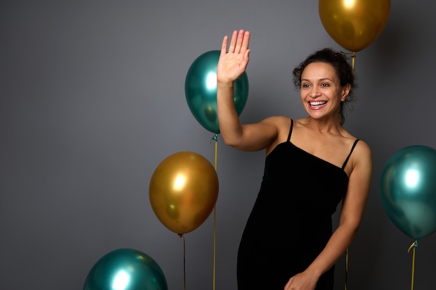 Mujer bonita alegre vestida con un elegante vestido de noche negro se encuentra sobre un fondo de pared gris con globos inflados metálicos dorados y verdes, saluda con la mano, sonríe con una sonrisa dentuda