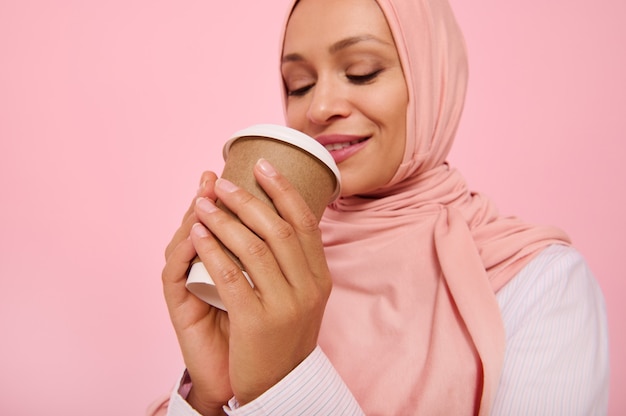 Mujer bonita árabe musulmana con la cabeza cubierta en hijab bebiendo bebida caliente, té o café de una taza de cartón desechable para llevar, de pie tres cuartas partes contra el fondo de color, copie el espacio. De cerca