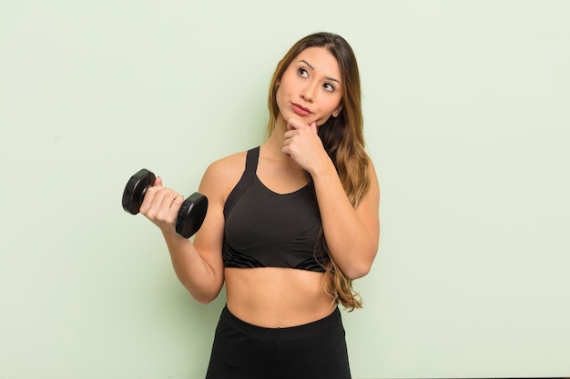 Mujer bonita asiática pensando, sintiéndose dudosa y confundida. concepto de fitness con una mancuerna