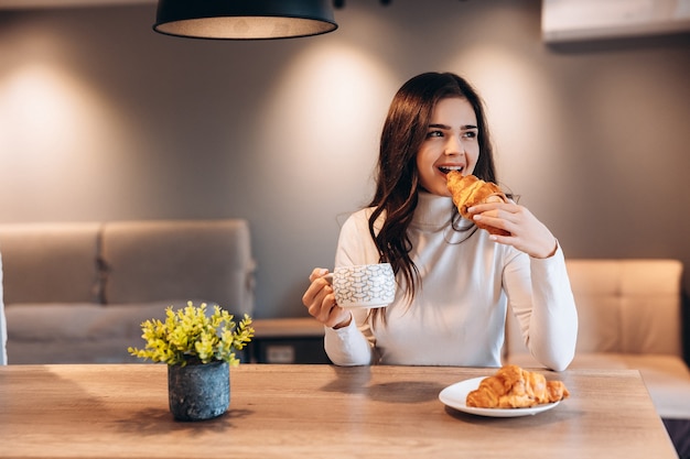 Foto mujer bonita con cabello negro brillante tomando café durante el desayuno. retrato interior de linda chica morena comiendo croissant y disfrutando del té en la mañana.