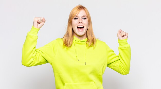 Mujer bonita cabeza roja gritando triunfalmente, con aspecto de ganador emocionado, feliz y sorprendido, celebrando