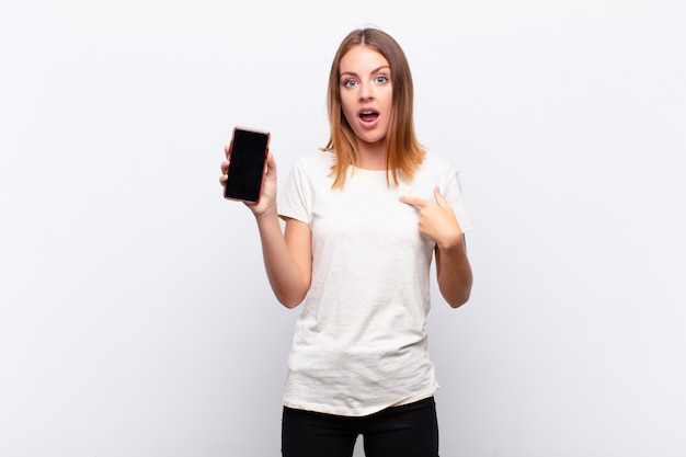 Mujer bonita cabeza roja mirando sorprendido y sorprendido con la boca abierta, apuntando a sí mismo sosteniendo un teléfono inteligente