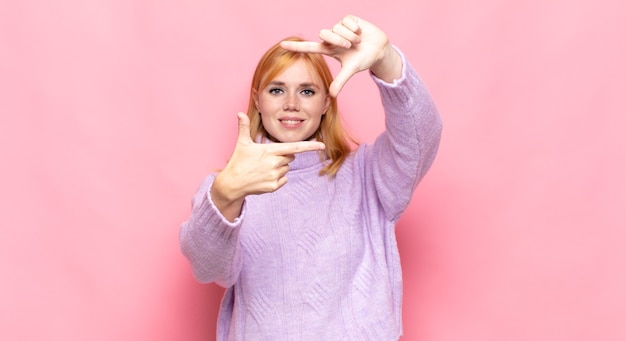 Mujer bonita cabeza roja que se siente feliz, amigable y positiva, sonriendo y haciendo un retrato o marco de fotos con las manos