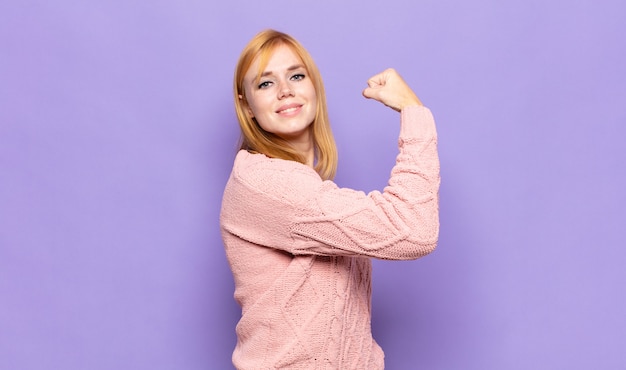 Mujer bonita cabeza roja que se siente feliz, satisfecha y poderosa, flexionando el ajuste y bíceps musculosos, luciendo fuerte después del gimnasio