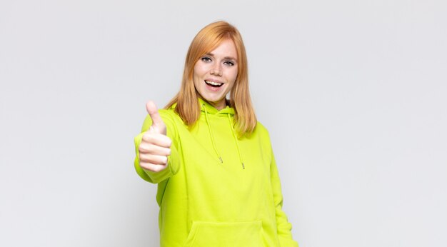 Mujer bonita cabeza roja que se siente orgullosa, despreocupada, segura y feliz, sonriendo positivamente con los pulgares hacia arriba