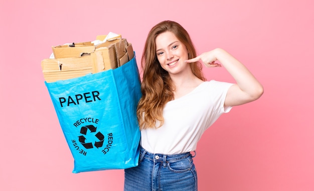 Mujer bonita cabeza roja sonriendo con confianza apuntando a su propia sonrisa amplia y sosteniendo una bolsa de papel reciclado