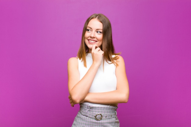 Mujer bonita cabeza roja sonriendo con una expresión feliz y segura con la mano en la barbilla, preguntándose y mirando a un lado contra la pared púrpura