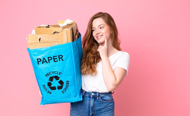 Mujer bonita cabeza roja sonriendo con una expresión feliz y segura con la mano en la barbilla y sosteniendo una bolsa de papel reciclado