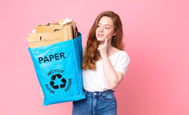 Mujer bonita cabeza roja sonriendo felizmente y soñando despierto o dudando y sosteniendo una bolsa de papel reciclado