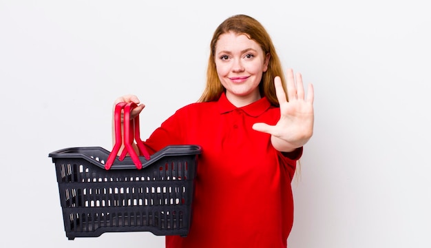 Foto mujer bonita de cabeza roja sonriendo y luciendo amigable mostrando el concepto de cesta de compras vacía número cinco