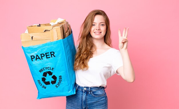 Mujer bonita cabeza roja sonriendo y mirando amigable, mostrando el número tres y sosteniendo una bolsa de papel reciclado