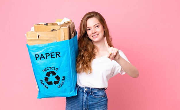 Mujer bonita cabeza roja sonriendo con orgullo y confianza haciendo el número uno y sosteniendo una bolsa de papel reciclado