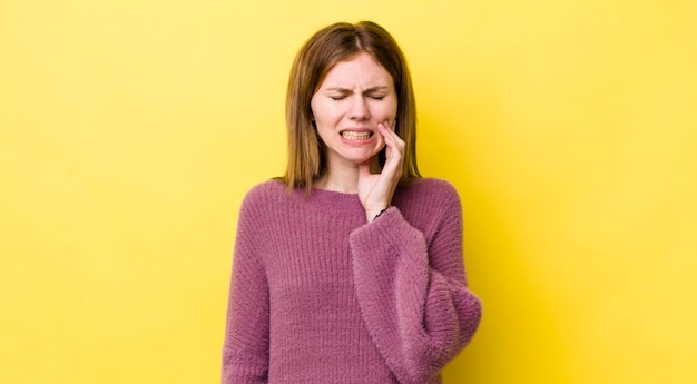 Mujer bonita de cabeza roja sosteniendo la mejilla y sufriendo dolor de muelas dolorosa sintiéndose enferma miserable e infeliz buscando un dentista