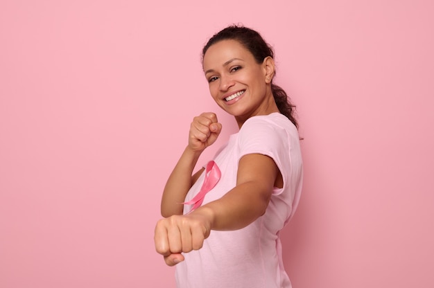 Mujer bonita con camiseta rosa y cinta de concienciación sobre el cáncer se encuentra en posición de combate para marcar la lucha contra el cáncer, en honor al 1 de octubre, sonríe mirando a la cámara, fondo de color, espacio de copia