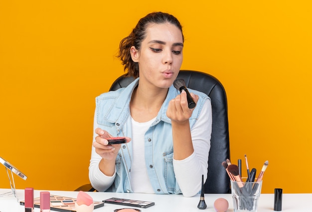 Mujer bonita caucásica complacida sentada a la mesa con herramientas de maquillaje sosteniendo rubor y soplando en pincel de maquillaje aislado en la pared naranja con espacio de copia