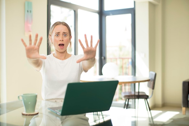 Mujer bonita caucásica con una computadora portátil sintiéndose estupefacta y asustada, temiendo algo aterrador, con las manos abiertas al frente diciendo: manténgase alejado