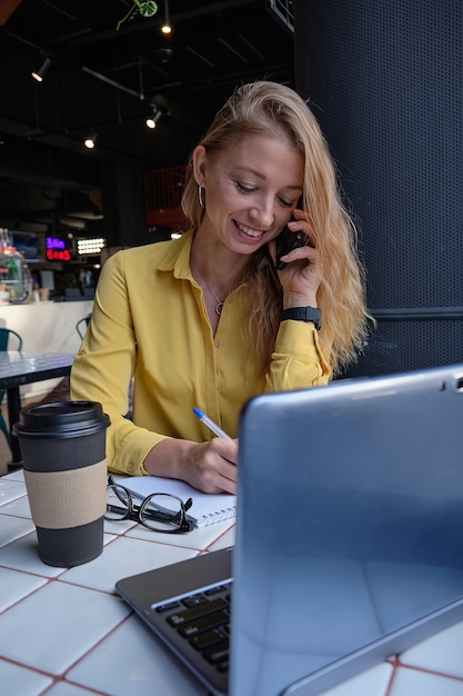 Mujer bonita caucásica joven sentada con el portátil en el café llamando por teléfono inteligente