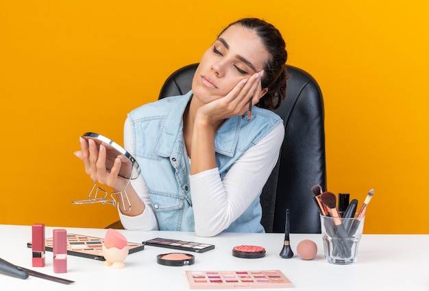 Mujer bonita caucásica soñolienta sentada a la mesa con herramientas de maquillaje poniendo la mano en la cara con pincel de maquillaje y espejo