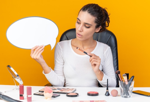 Foto mujer bonita caucásica sorprendida sentada en la mesa con herramientas de maquillaje sosteniendo pincel de maquillaje y mirando el bocadillo de diálogo