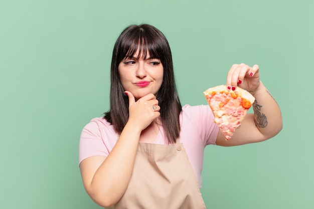 Mujer bonita chef dudando o expresión incierta y sosteniendo una pizza