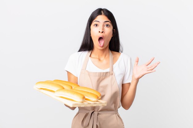 Mujer bonita chef hispana asombrada, conmocionada y asombrada con una sorpresa increíble y sosteniendo una bandeja con bollos de pan