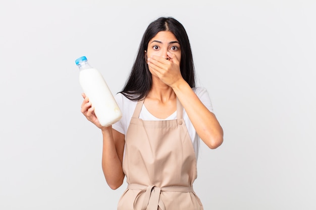 Mujer bonita chef hispana cubriendo la boca con las manos con un sorprendido y sosteniendo una botella de leche