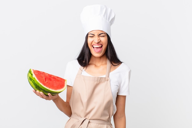 Mujer bonita chef hispana gritando agresivamente, mirando muy enojado y sosteniendo una sandía