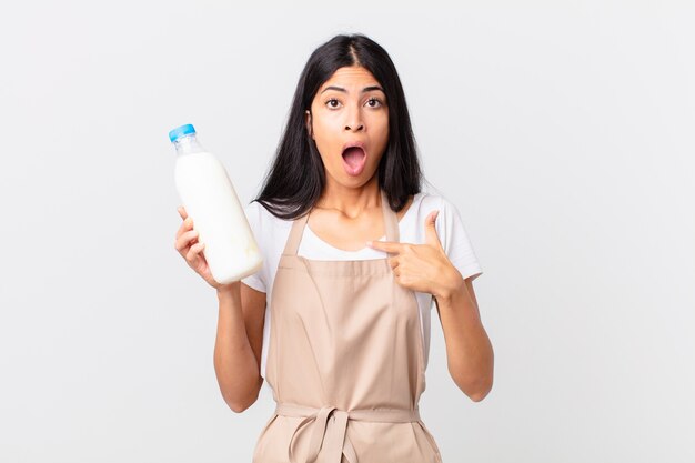 Mujer bonita chef hispana mirando conmocionado y sorprendido con la boca abierta, apuntando a sí mismo y sosteniendo una botella de leche