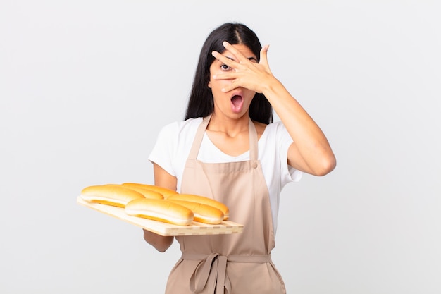 Mujer bonita chef hispana que parece sorprendida, asustada o aterrorizada, cubriendo la cara con la mano y sosteniendo una bandeja con bollos de pan