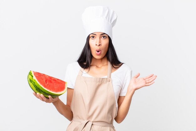 Mujer bonita chef hispana que parece sorprendida y conmocionada, con la mandíbula caída sosteniendo un objeto y sosteniendo una sandía