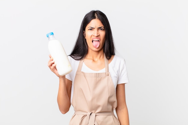 Mujer bonita chef hispana que se siente disgustada e irritada y con la lengua fuera y sosteniendo una botella de leche