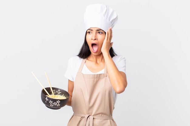 Mujer bonita chef hispana que se siente feliz, emocionada y sorprendida y sosteniendo un tazón de fideos