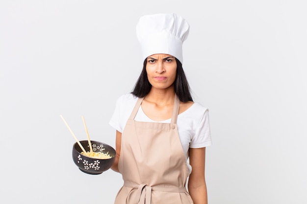 Mujer bonita chef hispana que se siente triste, molesta o enojada y mirando hacia un lado y sosteniendo un tazón de fideos