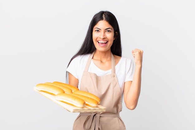 Mujer bonita chef hispana sintiéndose conmocionada, riendo y celebrando el éxito y sosteniendo una bandeja con bollos de pan