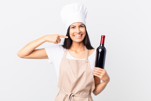 Mujer bonita chef hispana sonriendo con confianza apuntando a su propia sonrisa amplia y sosteniendo una botella de vino