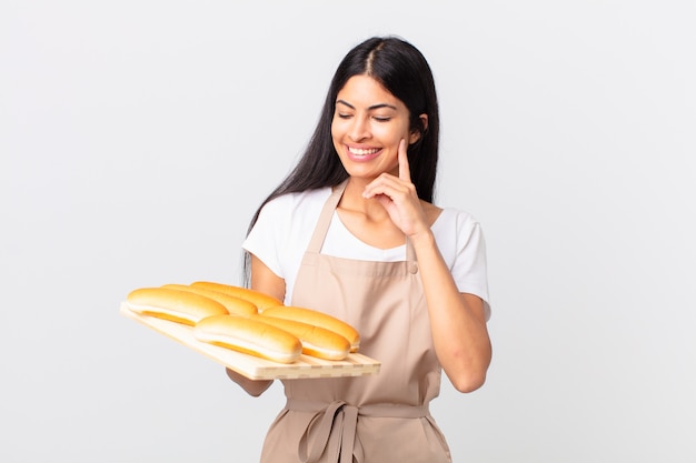 Mujer bonita chef hispana sonriendo felizmente y soñando despierto o dudando y sosteniendo una bandeja con bollos de pan