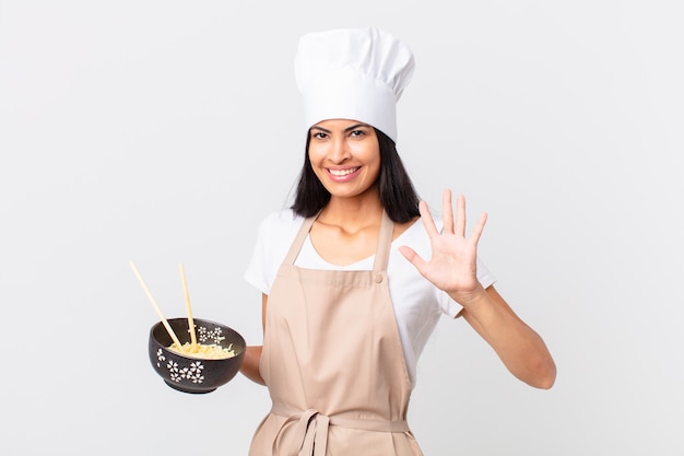 Mujer bonita chef hispana sonriendo y mirando amigable, mostrando el número cinco y sosteniendo un cuenco de fideos